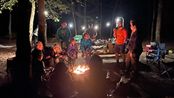 about 10 people sit or stand around a campfire roasting marshmallows